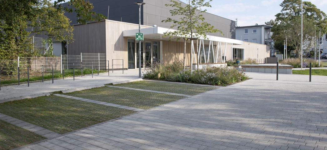 Modern exterior façade of a sports hall with anthracite-coloured panels in the upper area and wooden cladding in the lower area. In front of it is a forecourt paved with grey-lime, permeable concrete blocks with parking spaces made of grass pavers and shrubs, grasses and trees. 