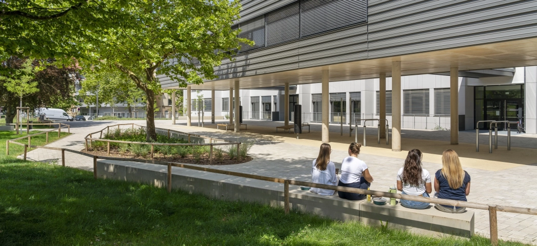 The schoolyard is surrounded by green meadows, trees and seating elements where children can chat with each other. A modern, multi-part school building can be seen in the background. The front part stands on concrete pillars and is clad in dark gray. The rear part has a white façade.