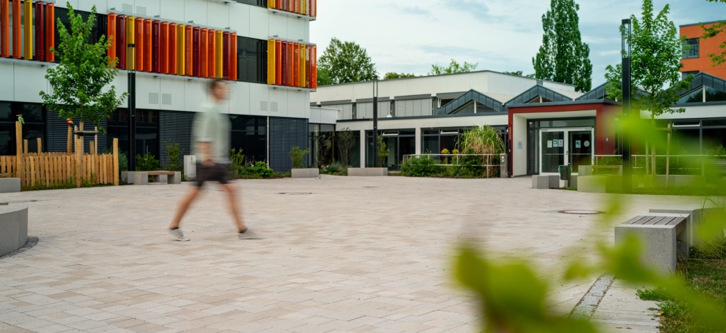Ein Mann geht über einen ansprechenden Schulhof mit vielen Sitzelementen und Grünflächen. im Vordergrund sind unscharf ein paar Zweige mit grünen Blättern zu erkennen. Im Hintergrund steht ein Schulgebäude mit weißen Fassaden und orange-gelben, geometrischen Verzierungen
