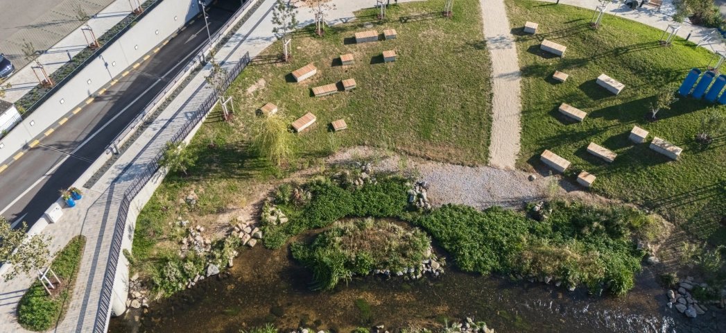 Aerial view of a newly designed park area with paved paths, a small stream, green areas with many benches and neighbouring play and traffic areas.