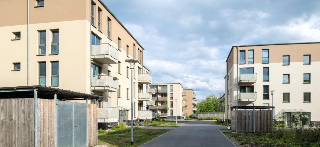 breite Durchfahrt zwischen mehreren gleichartigen Wohnblöcken mit beiger und brauner Fassade und Balkonen sowie Zugängen und kleinen Holzschuppen