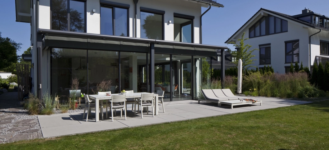 Modernes Einfamilienhaus mit Wintergarten und Glasfront vor heller Terrassenfläche mit Esstisch und Liegestuhl
