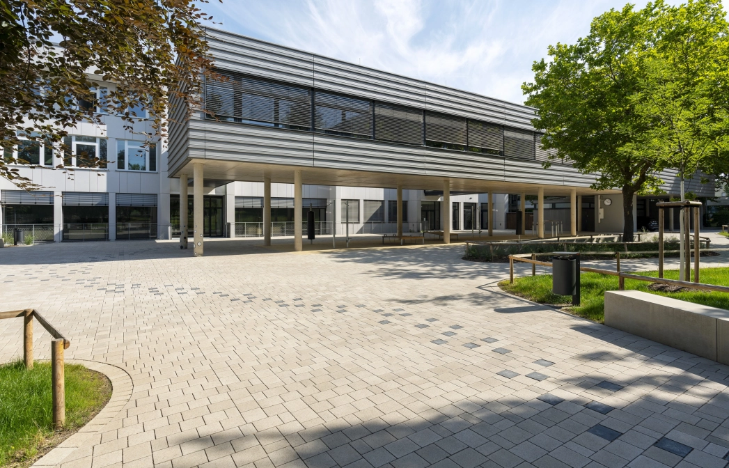 Open schoolyard surrounded by green meadows, trees and seating elements. A path consisting of anthracite-colored concrete blocks runs from right to left through the paved courtyard to a modern, multi-part school building. The front part stands on concrete columns and is clad in dark gray. The rear part has a white façade.
