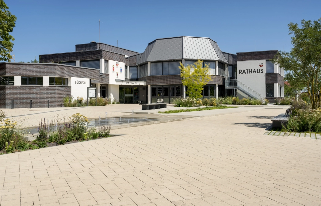 Open square in an urban environment. A water feature with fountain elements is placed in the middle of the paved square, surrounded by flowerbeds and trees. A modern town hall can be seen in the background, surrounded by the square.