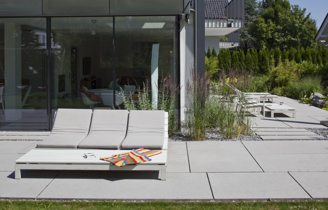Terrasse mit großformatigen grauen Terrassenplatten und breitem Liegestuhl vor Glasfront eines modernen Wohngebäudes