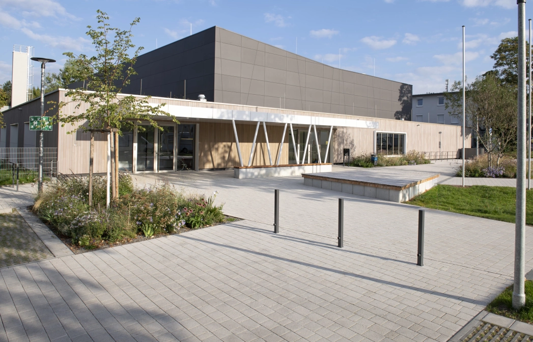 Modern exterior façade of a sports hall with anthracite-coloured panels in the upper area and wooden cladding in the lower area. In front of this is a forecourt paved with lime-grey, permeable concrete blocks with shrubs, grasses and a small tree.