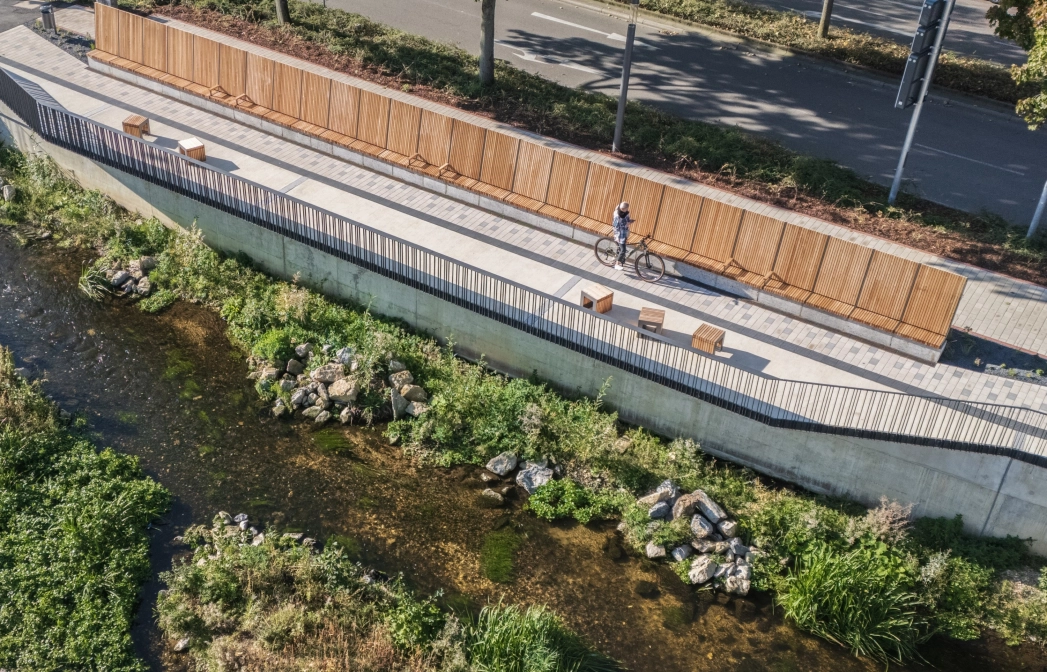 Aerial view of a riverside path along a shallow stream, lined with wooden benches and long wooden panelling. The path is paved with light-coloured concrete blocks, while a road with trees runs above it.