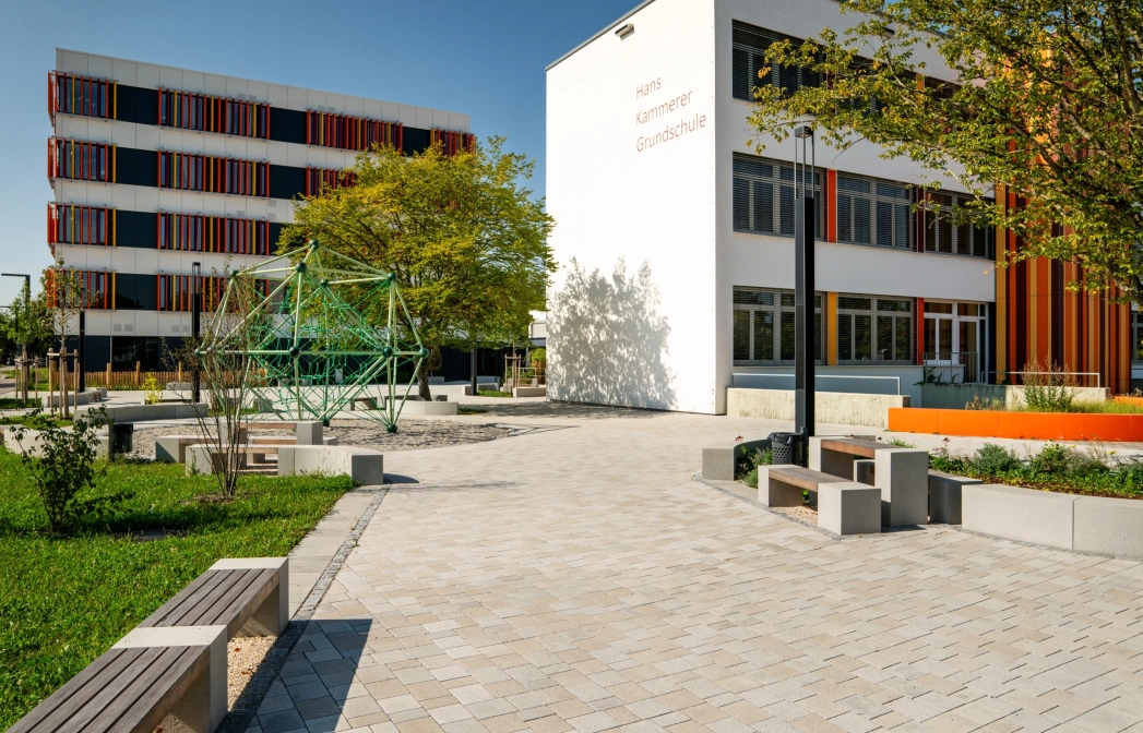 Ansprechender Schulhof mit vielen Sitzelementen und Grünflächen an einem sonnigen Tag mit blauem Himmel. Ein Klettergerüst innerhalb einer Sandfläche ziehrt den Hof und große Bäume spenden Schatten. Im Hintergrund stehen zwei Schulgebäude mit weißen Fassaden und orange-gelben, geometrischen Verzierungen