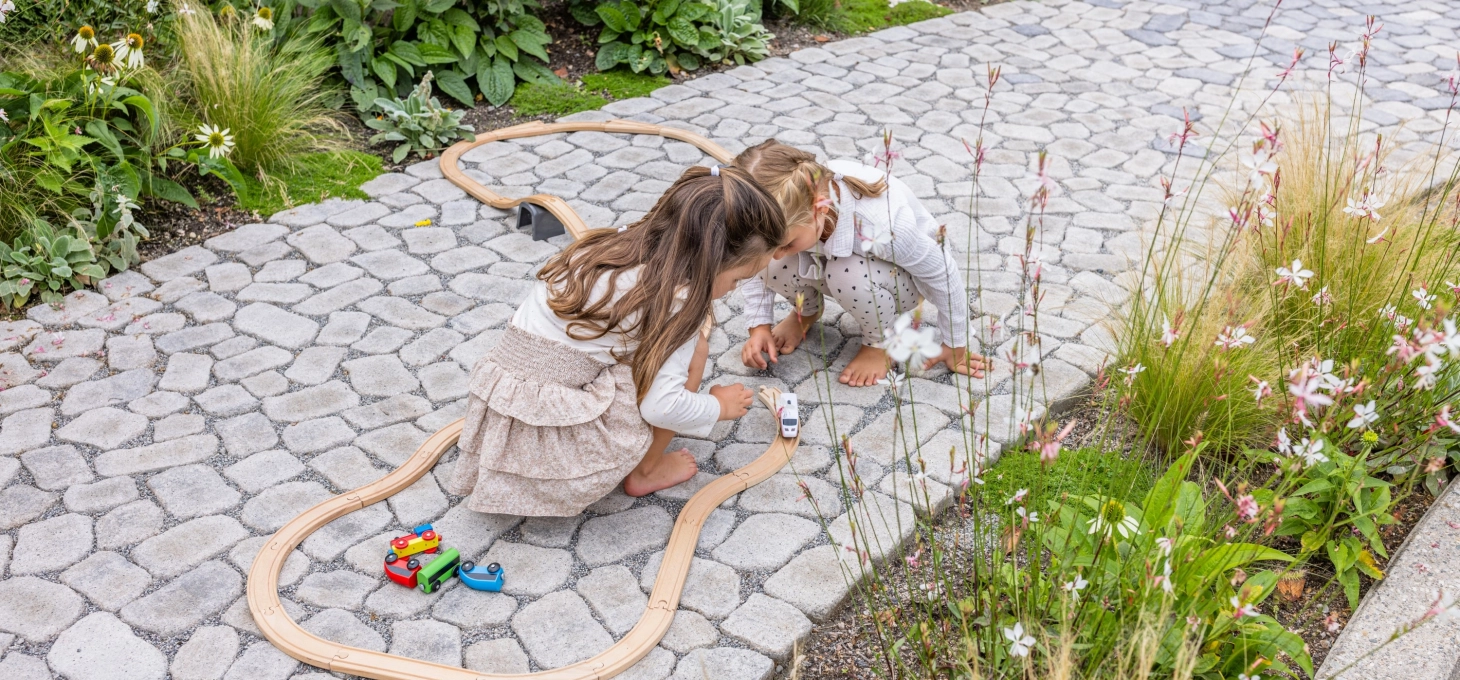 Zwei Mädchen spielen auf einem Gehweg aus kleinen, rundlichen Pflastersteinen GDM.Bolero stone  mit Holzfahrzeugen 