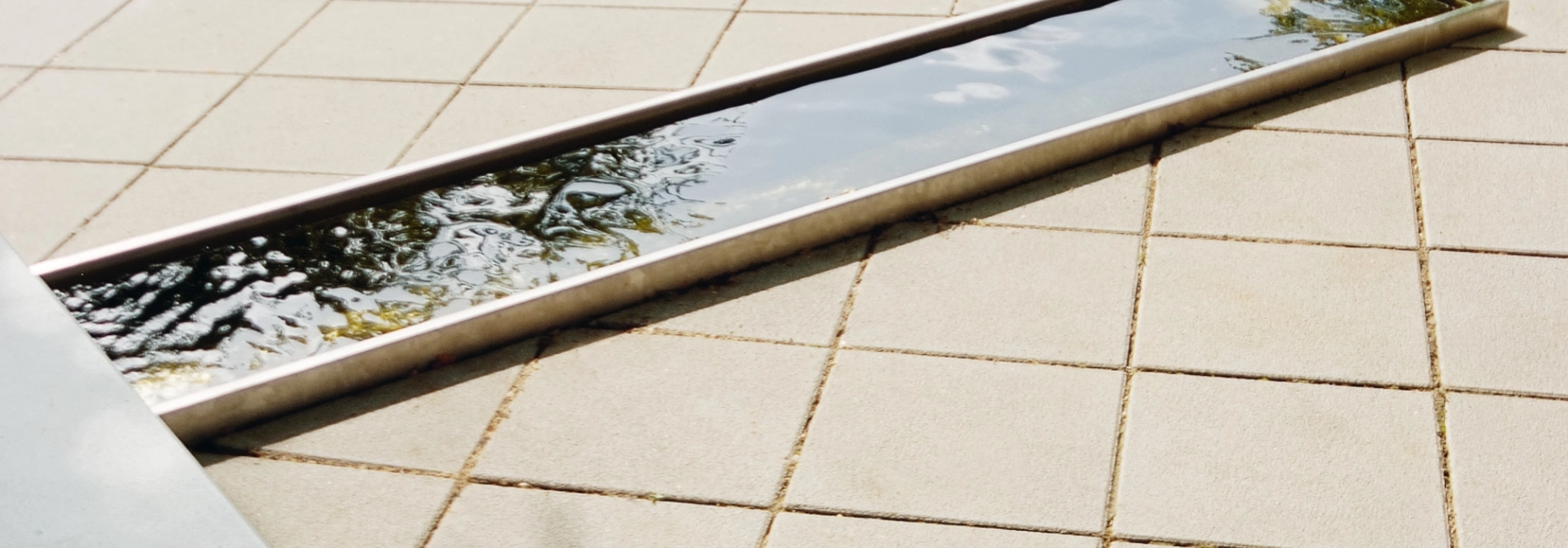 sandfarben gepflasterter Platz in Parkanlage mit geometrisch angelegtem Wasserlauf hin zu quadratischen Sitzblöcken