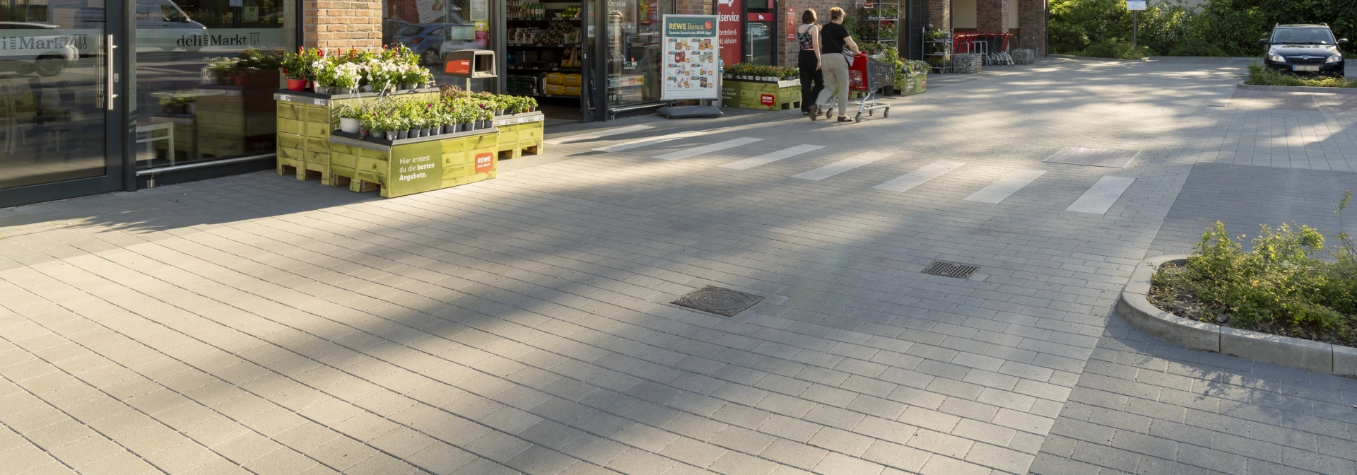GDM.KLIMASTEIN aBG vor dem Eingangsbereich des REWE Markts Christian Schulz mit Zebrastreifen und Backsteinfassade.