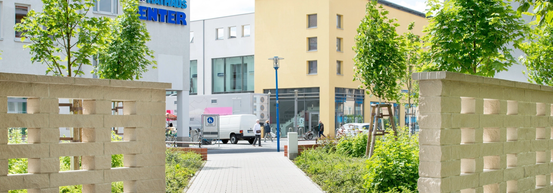 sandfarbene Betonsteinmauer mit fensterähnlichen Aussparungen rechts und links von einem Fußgängerweg in Richtung Innenstadt