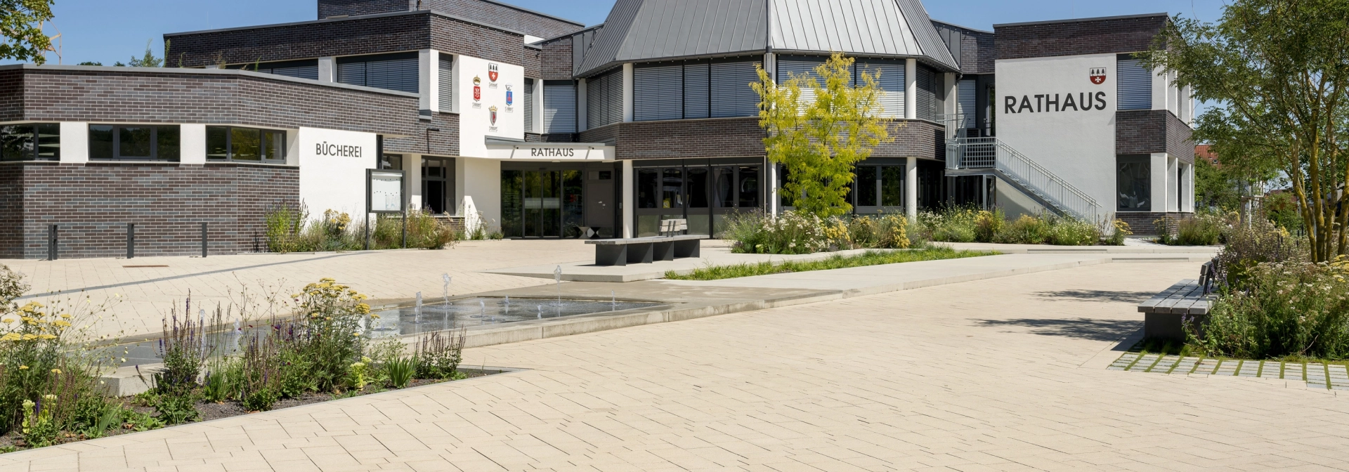 Open square in an urban environment. A water feature with fountain elements is placed in the middle of the paved square, surrounded by flowerbeds and trees. A modern town hall can be seen in the background, surrounded by the square.