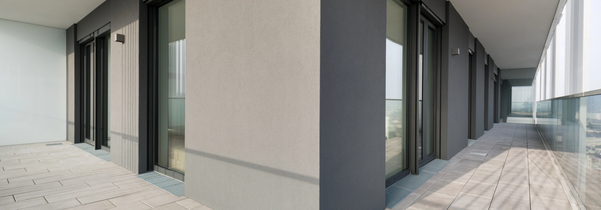 seitlicher Balkon mit hellen Terrassenplatten neben grau-brauner Fassade mit mehreren hohen Fenstern und Türen