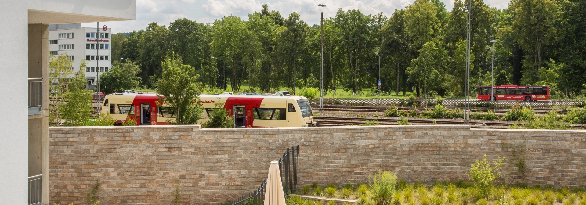 helle sandfarbene Schallschutzwand in einem begrünten Garten mit dahinter liegenden Schienenverkehr