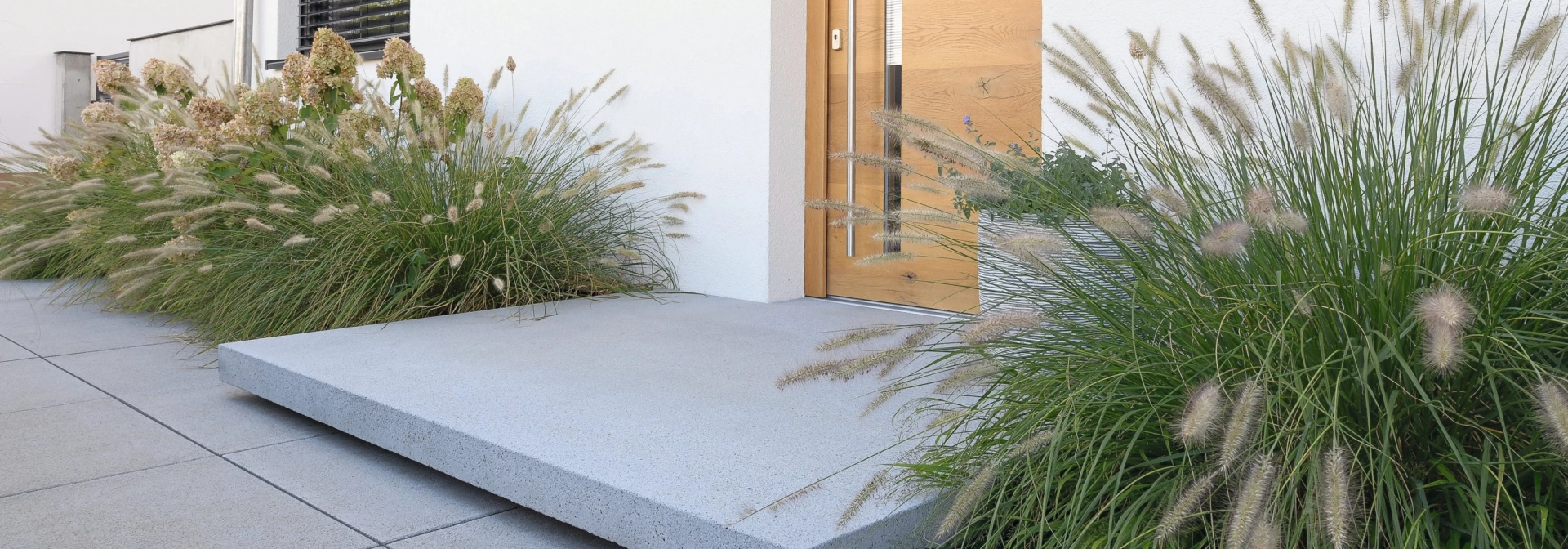 Moderner Hauseingang mit grauem Betonpodest und heller Haustüre aus Holz. Davor sind graue Betonsteinplatten verlegt. Links und Rechts von der Haustüre sind große, grüne Sträucher.