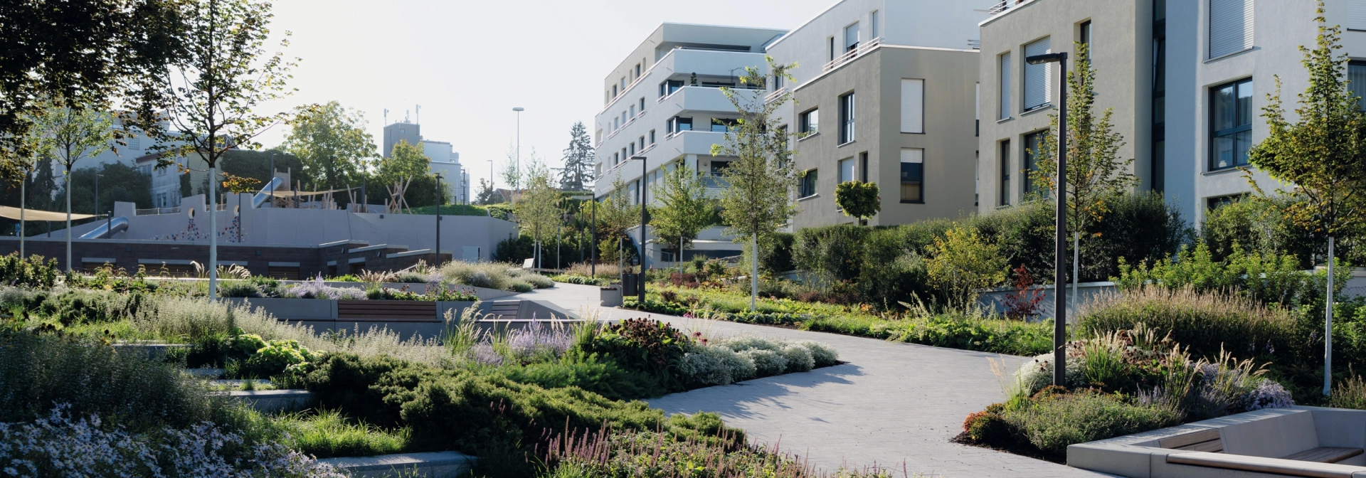 Ein grau gepflasterter Weg, welcher an modernen Wohngebäuden vorbeiführt. Der Weg ist seitlich von vielen grünen Pflanzen umgeben. Im Vordergrund sind Stufen, welche ebenfalls von vielen Sträuchern und Gräsern umgeben ist.