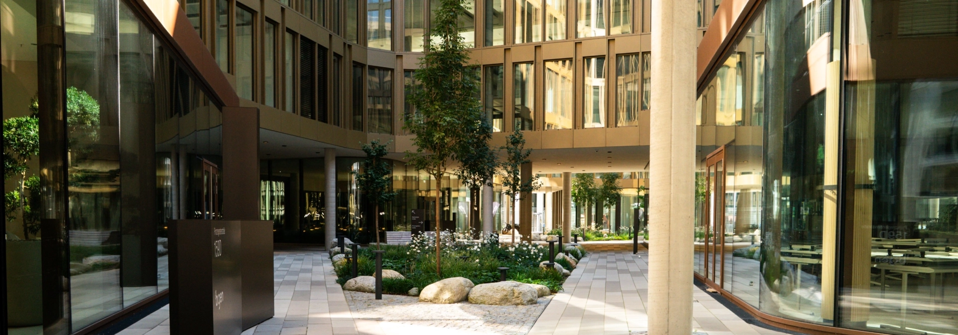 Innenhof eines großen Bürokomplexes mit Fensterfassade. Die Fläche ist mit dem kalksteinfarbenem GDM.SCENIC individual gepflastert. Außerdem findet man eine hohe beige Säule, ein Beet mit Sträuchern und kleinen Bäumen und eine schwarze Tafel.