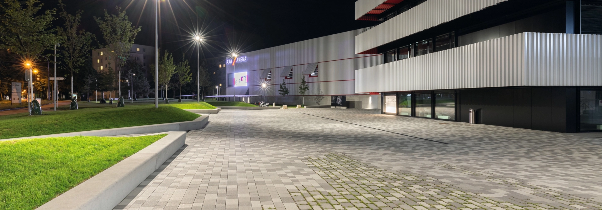 Gepflasterter Platz mit teilweise begrünter Fuge und an Grünfläche angrenzende Sitzmauern vor einem Sportzentrum