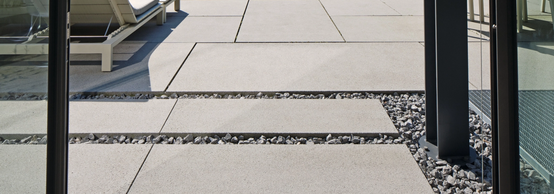 großformatige hellgraue Terrassenplatten und Sonnenliege hinter Schiebetür aus Glas mit schwarzem Türrahmen