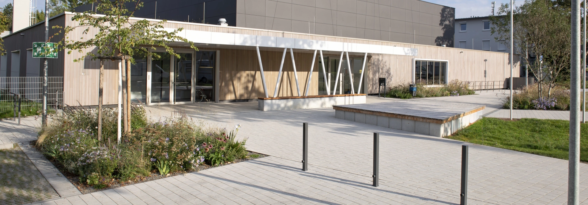 Modern exterior façade of a sports hall with anthracite-coloured panels in the upper area and wooden cladding in the lower area. In front of this is a forecourt paved with lime-grey, permeable concrete blocks with shrubs, grasses and a small tree.