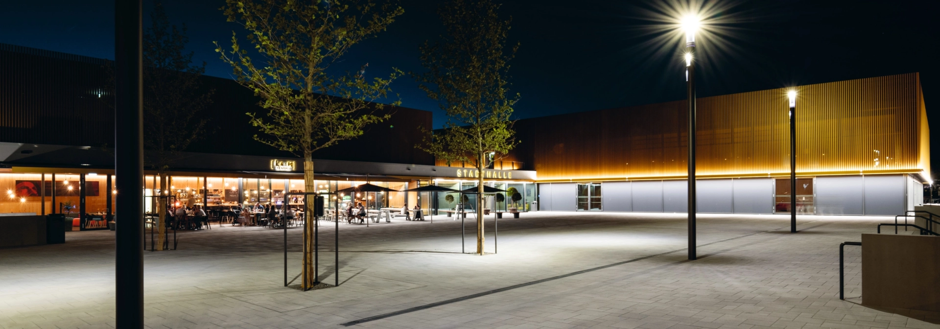 Beleuchteter, gepflasterter Platz vor Stadthalle bei Nacht mit Baumbepflanzung