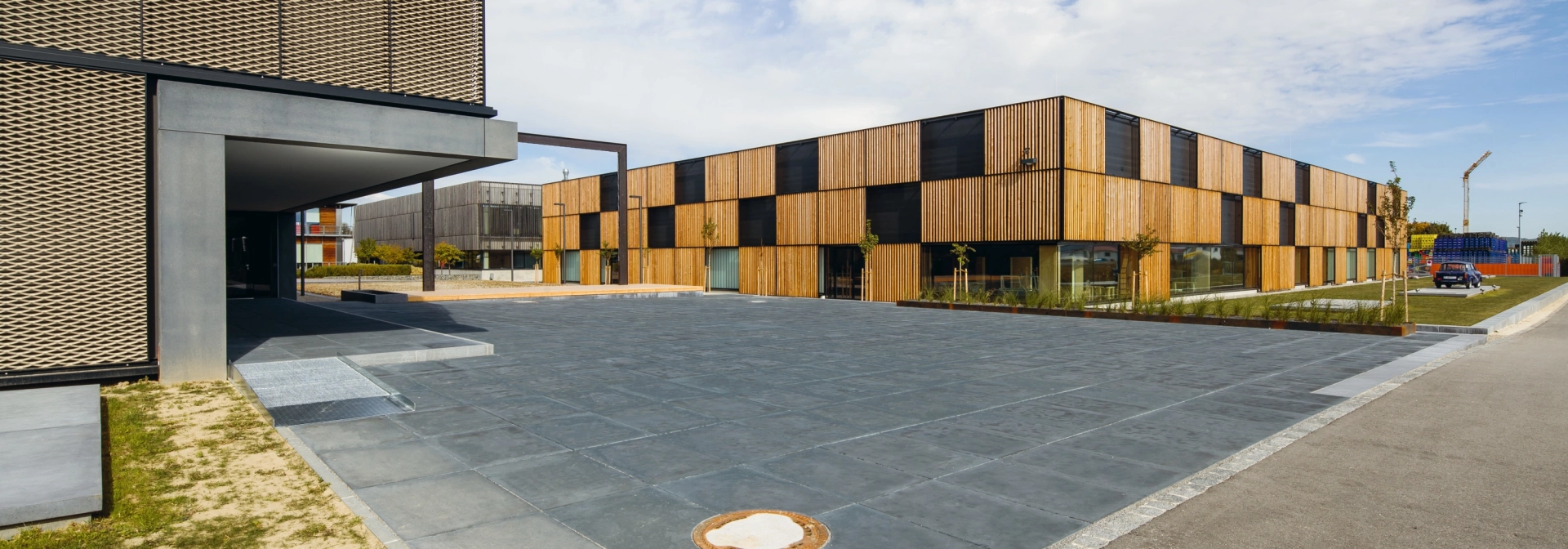 großer dunkel gepflasterter Platz vor modernem Firmengebäude mit Holzfassade und Flachdach