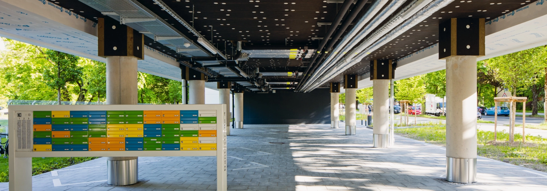 offener Eingangsbereich unter Gebäude mit schwarzer Decke und weißen Säulen mit bunten Briefkästen