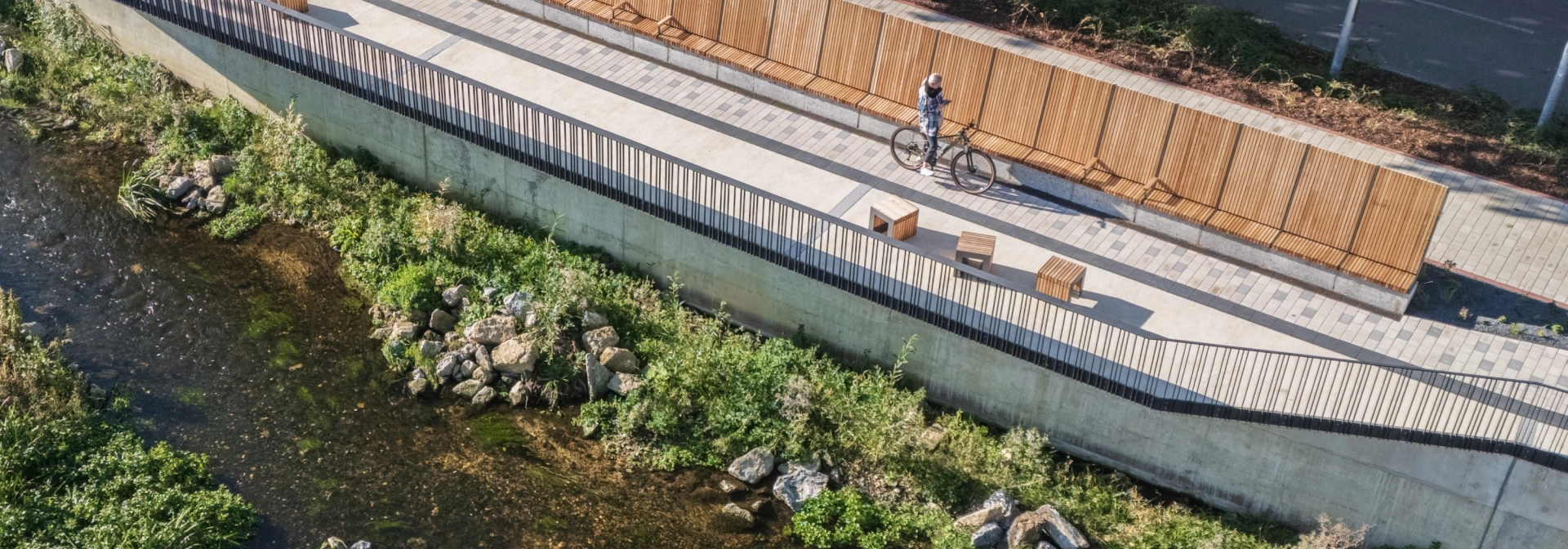 Aerial view of a riverside path along a shallow stream, lined with wooden benches and long wooden panelling. The path is paved with light-coloured concrete blocks, while a road with trees runs above it.