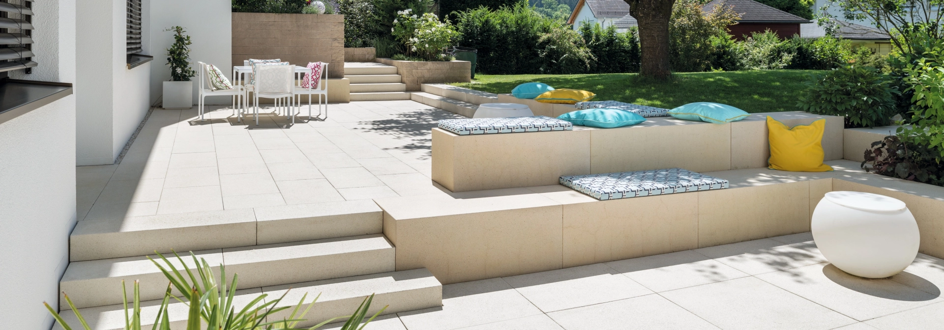 Patio slabs in a landscaped garden with a large tree and concrete seating elements.