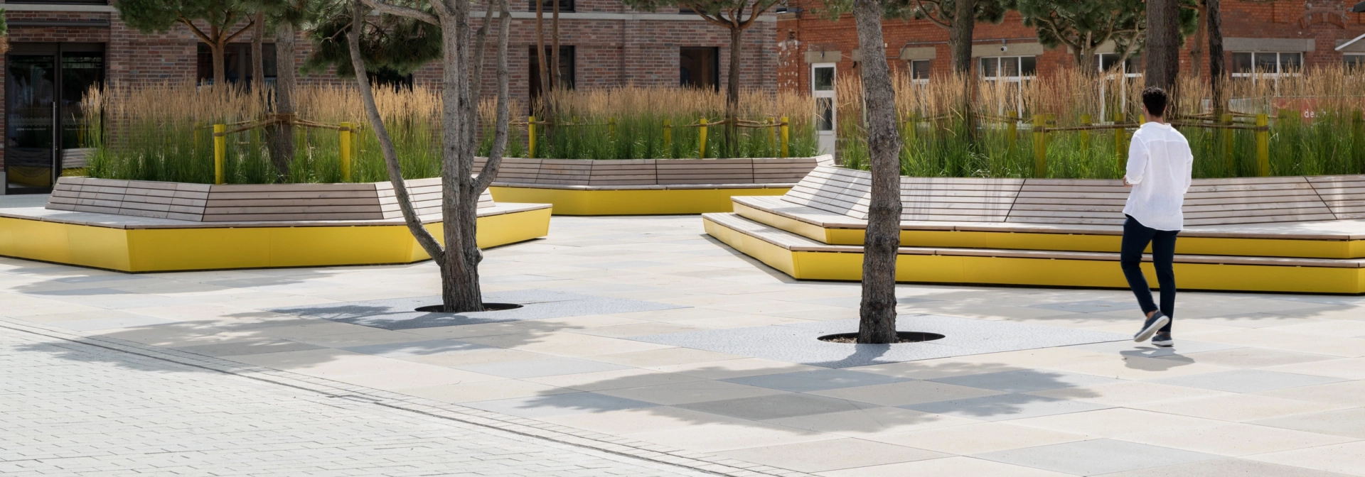 GDM.SCENIC individual auf einem großzügigen Platz mit Bäumen, gelben Sitzbänken und bepflanzten Aufenthaltsbereichen vor Backsteinarchitektur.