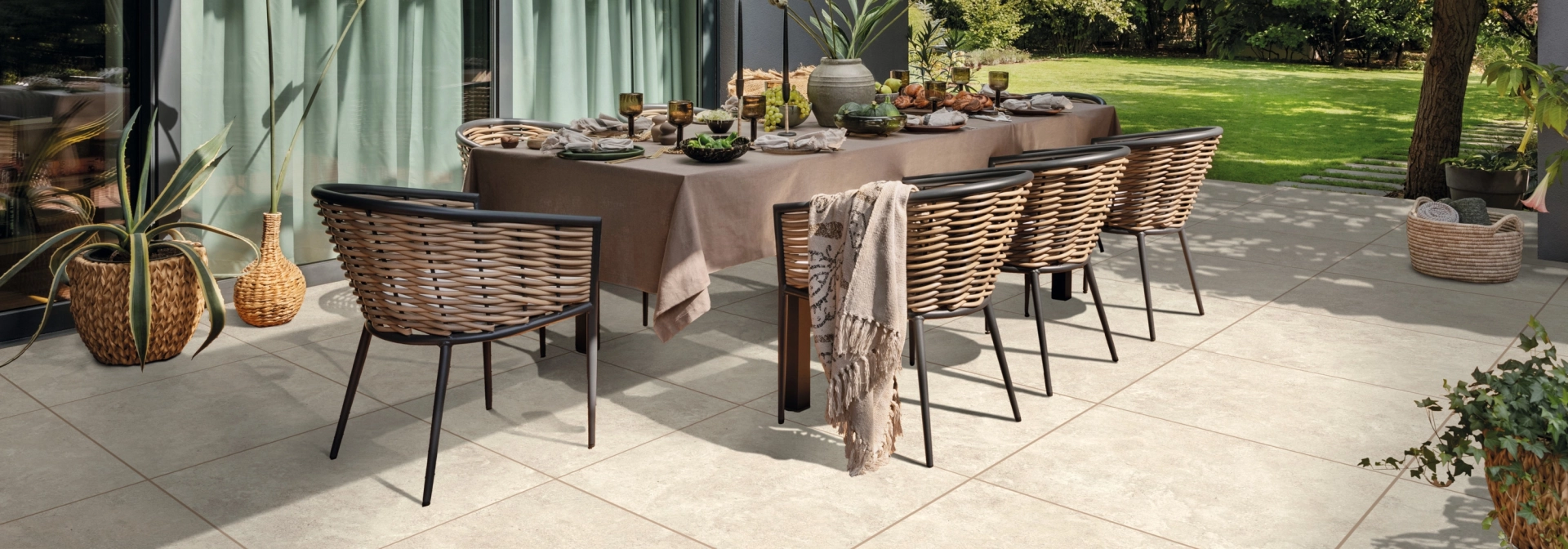 Moderne Terrasse mit schön gedecktem und dekorierten Tisch mit vielen Stühlen und Keramik-Verbundplatten am Boden verlegt. Im Hintergrund ist ein grüner Garten und das Wohnhaus mit großen Fenstern und anthrazit-farbener Fassade zu erkennen.