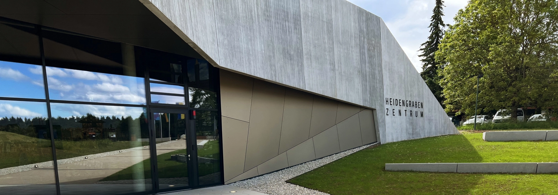 Im Vordergrund sind fünfeckige, kalksteinfarbene Betonsteine verlegt. Dahinter ist eine moderne Gebäudefassade in Holzoptik mit Fensterfront. Daneben ist eine grüne Rasenfläche.