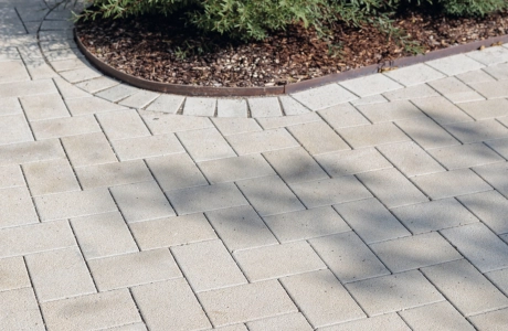 Close-up of a path made of beige GDM.KLIMASTEIN, bordering a circular flowerbed with dense greenery and dark mulch. In the upper area, shadows from surrounding elements fall onto the paving.