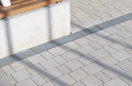 Close-up of grey-lime, permeable concrete paving stones - the GDM.KLIMASTEIN. Next to it is a concrete bench with wooden slats as a seat. Two white supports end in it