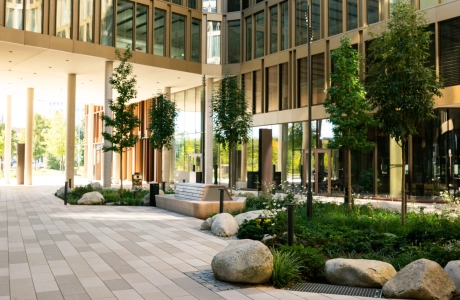 Innenhof eines großen Bürokomplexes mit Fensterfassade. Die Hoffläche ist mit dem kalksteinfarbenem GDM.SCENIC individual gepflastert. Außerdem findet man Beete mit Sträuchern, kleinen Bäumen und Steinen.