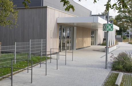 Roofed entrance area of a modern sports hall with a façade made of anthracite-coloured panels and wood. In front of the building there is a forecourt paved with permeable, grey-lime concrete blocks with bicycle parking racks, green areas and a street lamp with a collection point sign.