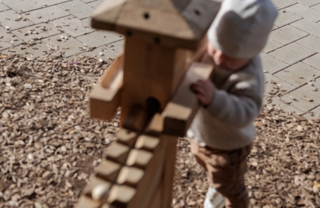A child is playing on a wooden marble run on a floor made of wood chips. Behind it is a path made of light-coloured concrete blocks. 