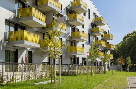 Wohngebäude mit heller Außenfassade und gelben Balkonbrüstungen. Davor ist ein Grünstreifen mit kleinen Bäumen und eine weitere Fläche, welche mit dem GDM.SCADA liner gepflastert ist. Zwischen den einzelnen Rasenfugensteinen wächst Gras.