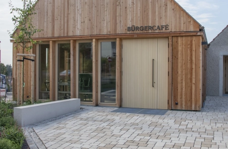 A modern building with a façade of vertical wooden slats, labelled "Bürgercafé". In front of the building is a large paved square with surrounding flowerbeds planted with flowers and shrubs. The sky is clear and slightly cloudy.