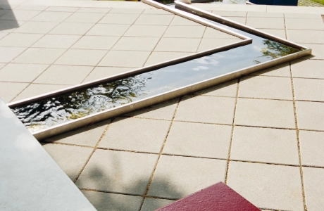 sandfarben gepflasterter Platz in Parkanlage mit geometrisch angelegtem Wasserlauf hin zu quadratischen Sitzblöcken
