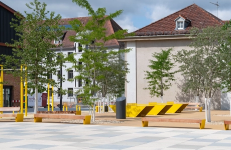 GDM.SCENIC individual auf einem großzügigen Platz mit gelben Sitzbänken, jungen Bäumen und offenen Aufenthaltsbereichen.