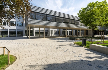 Open schoolyard surrounded by green meadows, trees and seating elements. A path consisting of anthracite-colored concrete blocks runs from right to left through the paved courtyard to a modern, multi-part school building. The front part stands on concrete columns and is clad in dark gray. The rear part has a white façade. 