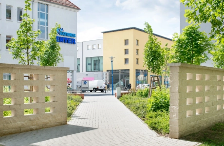 sandfarbene Betonsteinmauer mit fensterähnlichen Aussparungen rechts und links von einem Fußgängerweg in Richtung Innenstadt