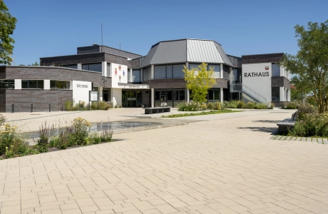 Open square in an urban environment. A water feature with fountain elements is placed in the middle of the paved square, surrounded by flowerbeds and trees. A modern town hall can be seen in the background, surrounded by the square.