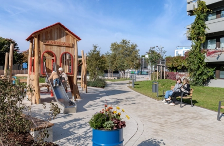 Playground area with a large wooden playhouse and a metal slide with two people on it. The surrounding path is made of beige paving stones and leads through a green area. On the right are two more people sitting on a wooden bench, in the foreground is a planted blue tub, and trees and modern buildings can be seen in the background.