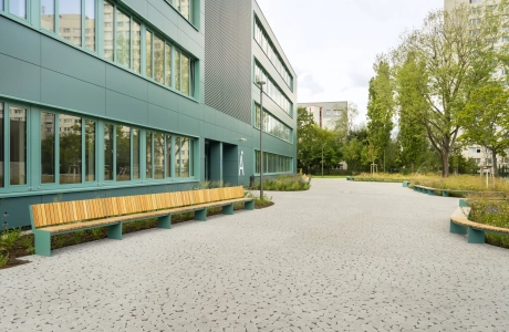 Ein mit rundlich und natürlich wirkenden Betonsteinen gepflasterter Schulhof, welcher rechts neben einem modernen Schulgebäude mit petrolfarbener Fassade liegt. Um den gepflasterten Bereich ist alles mit Streuchern, Wiesen und Bäumen bepflanzt, weche mit Siztbänken eingerahmt werden.