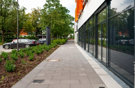 Ein anthrazit gepflasterter Geweg führt an einem auf der rechten Seite stehenden Gebäude vorbei. Das Gebäude hat eine große Glasfront, eine weiße Fassade und orange-gelben, geometrischen Verzierungen. Auf der linken Seite wird der Gehweg durch ein Beet mit Sträuchern von einem Parkplatz getrennt. 