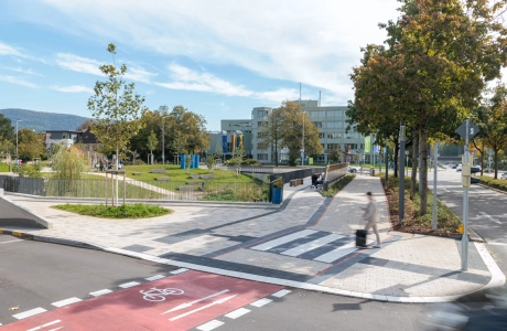 Overview of an urban pathway in which a wide footpath is predominantly paved with beige GDM.KLIMASTEIN. Red and white paving stones made of GDM.TETRAGO stone are integrated to mark the crossing area. The paths border a landscaped car park with lawn, trees, seating areas and a watercourse. A cycle path runs in the foreground.