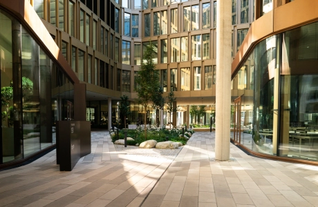 Innenhof eines großen Bürokomplexes mit Fensterfassade. Die Fläche ist mit dem kalksteinfarbenem GDM.SCENIC individual gepflastert. Außerdem findet man eine hohe beige Säule, ein Beet mit Sträuchern und kleinen Bäumen und eine schwarze Tafel.